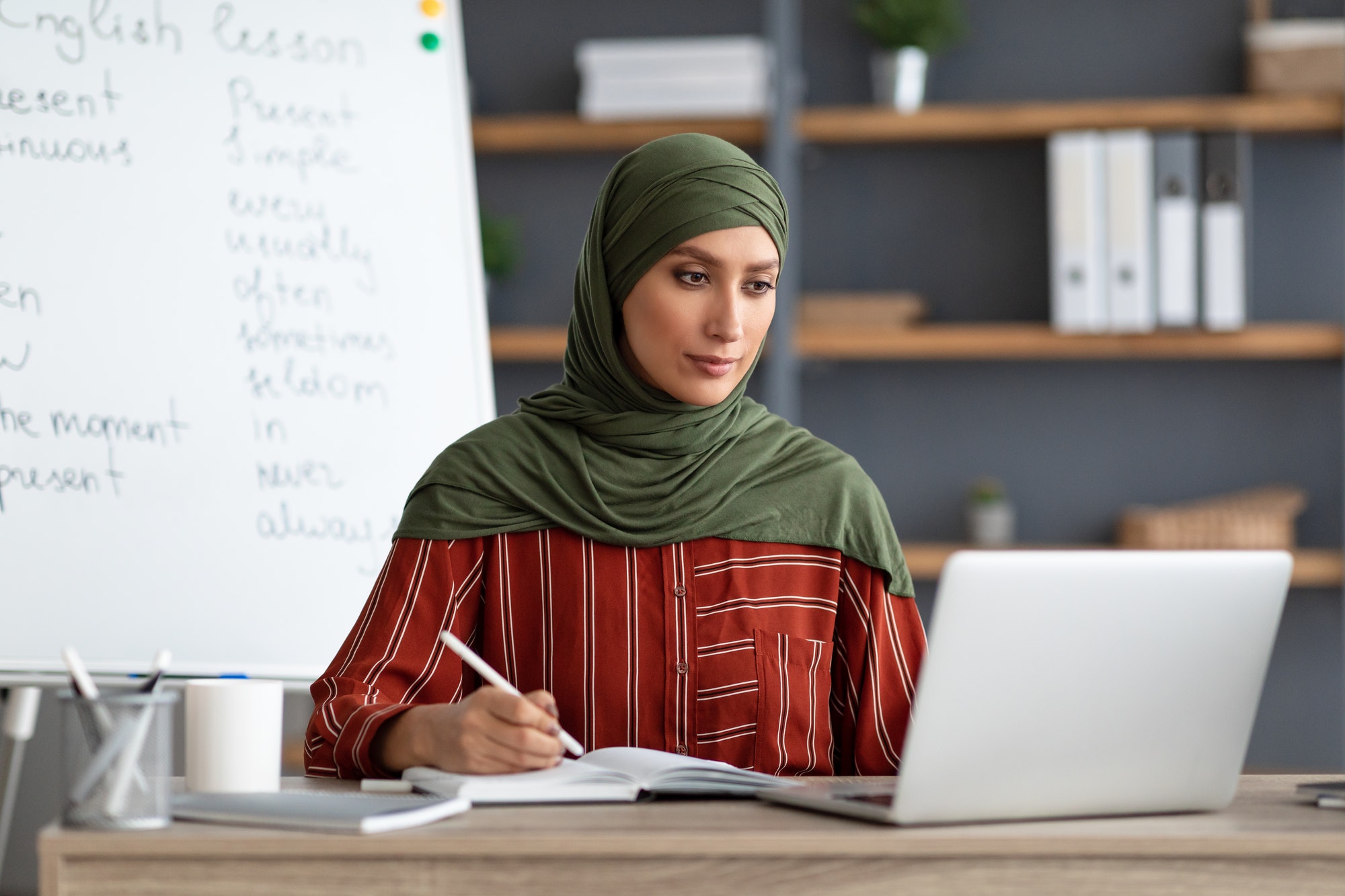 Islamic teacher in headscarf sitting at desk looking at laptop Islamic teacher in headscarf sitting at desk looking at laptop